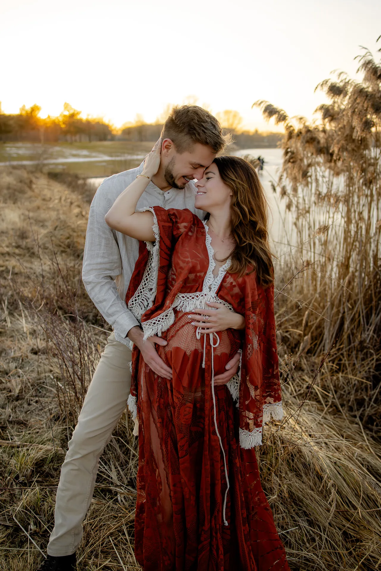 Schwangeres Paar kuschelt im roten Umstandskleid bei Sonnenuntergang in der Natur