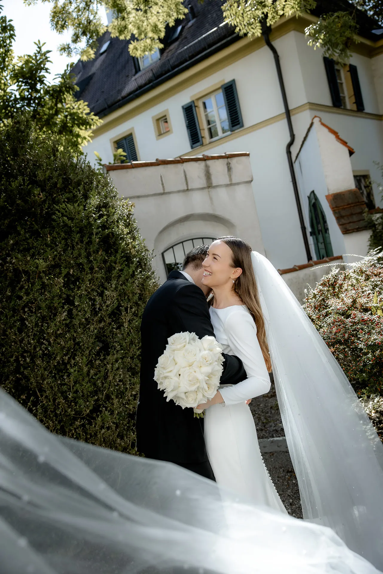 Brautpaar umarmt sich vor historischem Schlössl Asam mit wehendem Schleier