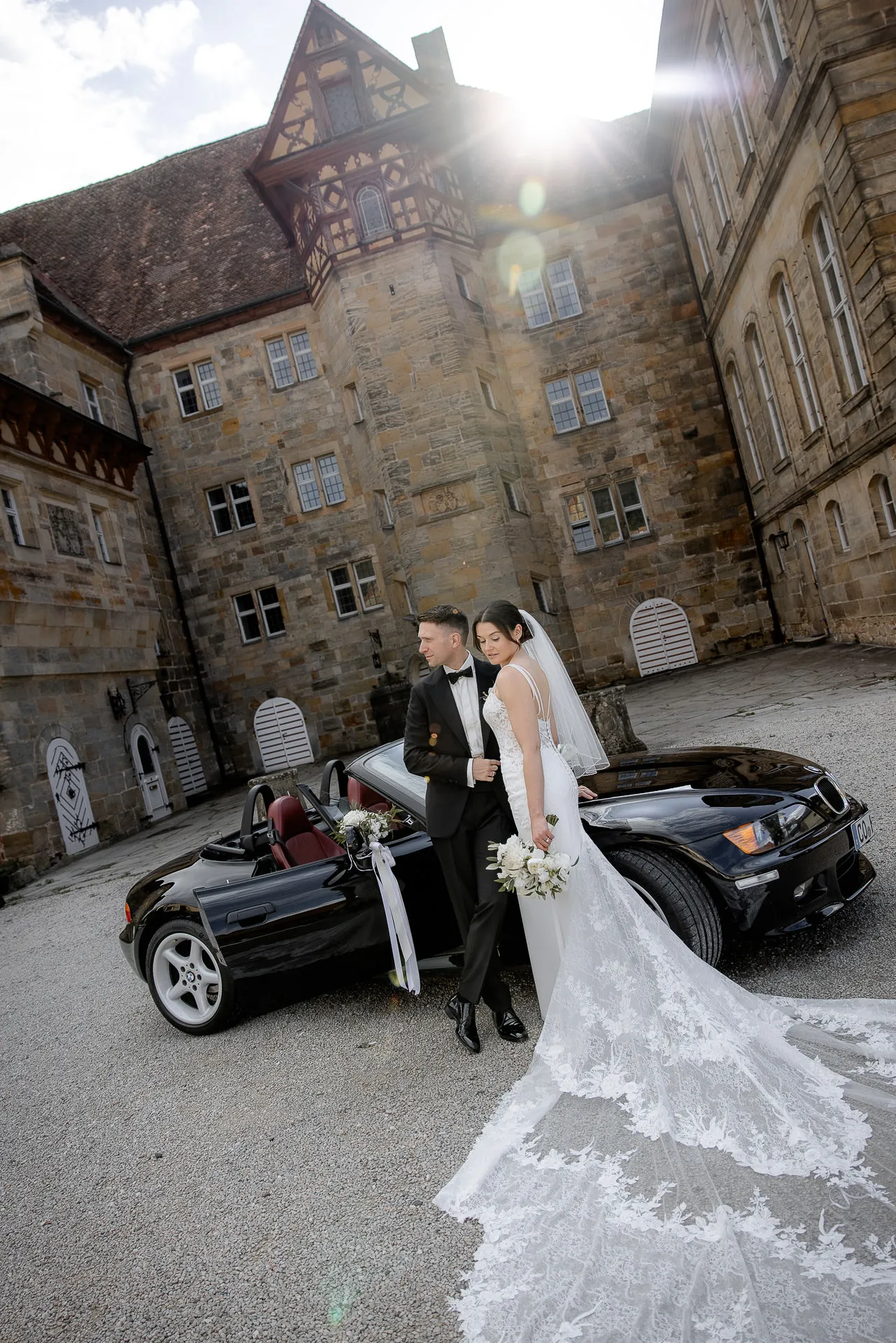 Elegantes Brautpaar mit Cabrio vor historischem Schloss Eyrichshof mit Sonnenstrahlen