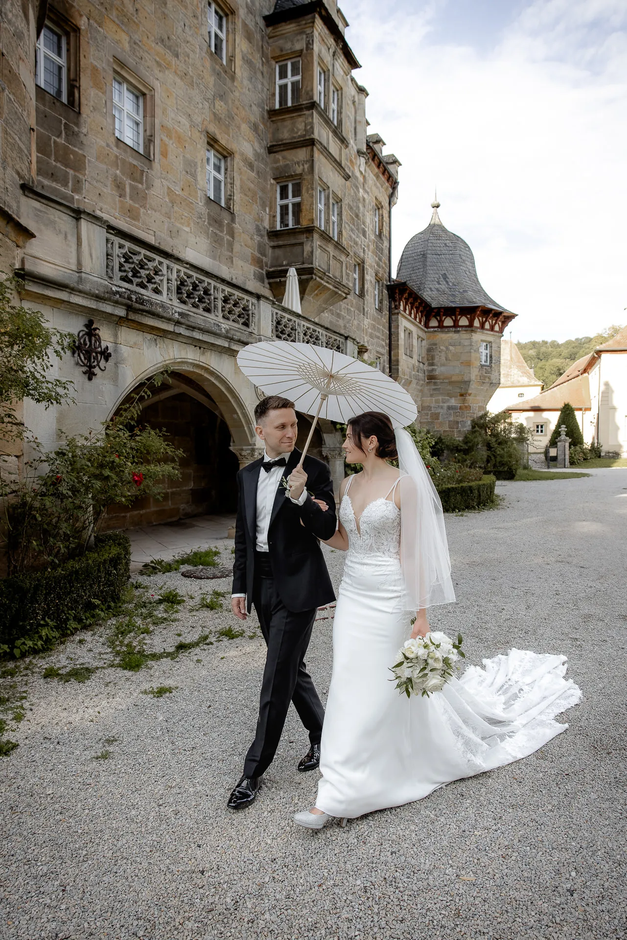 Brautpaar mit weißem Sonnenschirm spaziert durch Schlosshof Eyrichshof bei Hochzeit