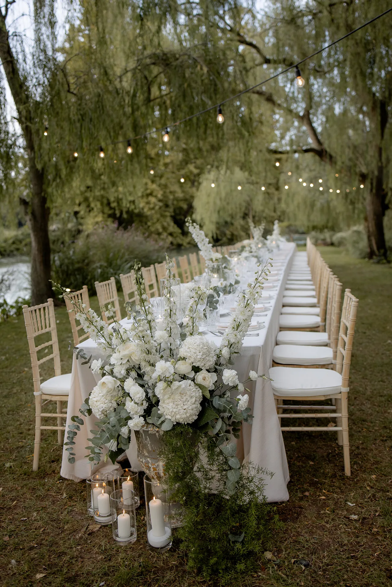 Romantische Abendhochzeit-Tafel mit weißer Blumendeko und Lichterketten unter Weiden